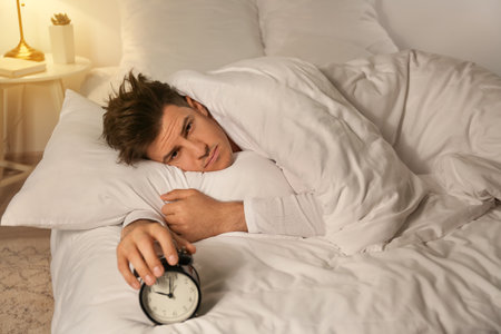 Young man with alarm clock suffering from insomnia in the bedroomの写真素材