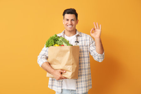 Young man with food in bag showing OK gesture on color backgroundの写真素材