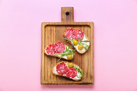 Wooden board of tasty sandwiches with cream cheese and salami on pink backgroundの写真素材
