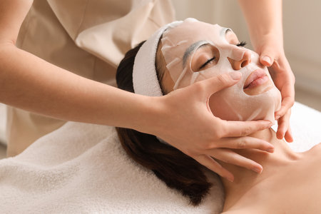 Cosmetologist applying sheet mask onto young woman's face in salon, closeupの写真素材