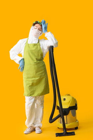 Tired housewife in protective costume and with vacuum cleaner on color backgroundの写真素材