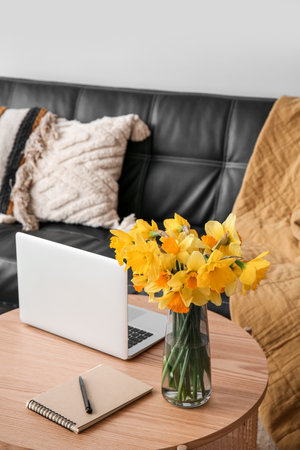 Modern laptop, vase with narcissus flowers and notebook on coffee table in living roomの写真素材