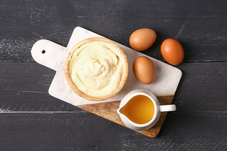 Bowl with tasty mayonnaise sauce and ingredients on dark wooden background