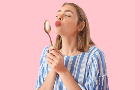 Young woman with golden spoon blowing kiss on pink backgroundの写真素材
