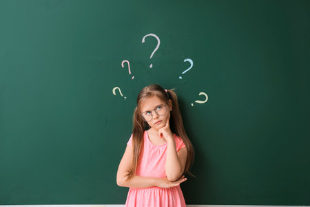 Thoughtful little schoolgirl near blackboard in classroomの写真素材