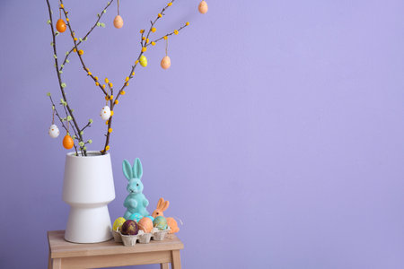 Vase with tree branches, Easter eggs and toy rabbits on stepladder near lilac wall