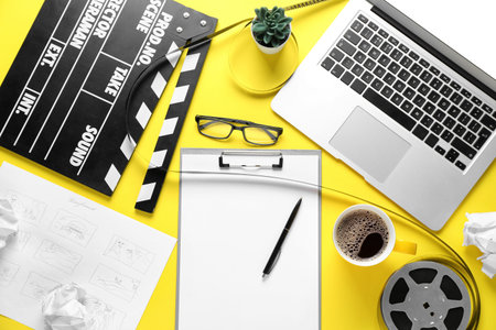 Clipboard with movie clapper, film reel, cup of coffee and laptop on yellow background