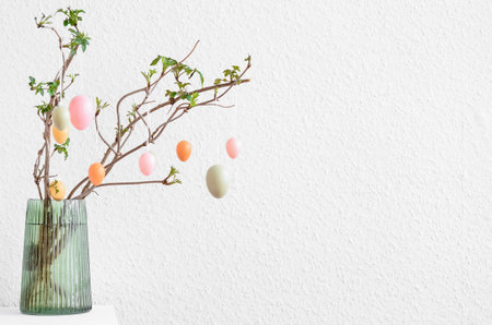 Vase with tree branches and Easter eggs on table near light wall