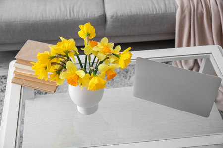 Modern laptop, vase with narcissus flowers and books on glass coffee table in living roomの写真素材