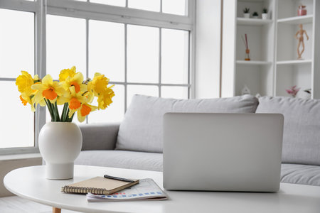 Modern laptop, vase with narcissus flowers, notebook and magazine on coffee table in living roomの写真素材