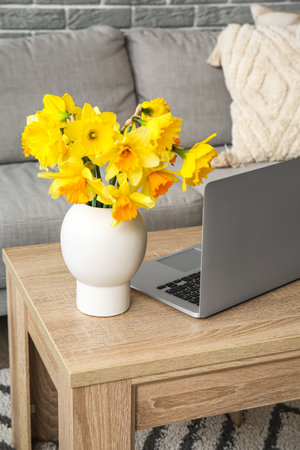 Modern laptop and vase with narcissus flowers on coffee table in living roomの写真素材