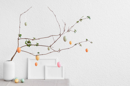 Vase with tree branch, Easter eggs and blank picture frame on table near light wallの写真素材