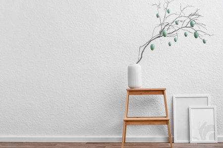 Vase with tree branch, Easter eggs and blank picture frames near light wall