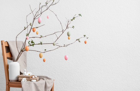 Vase with tree branches, Easter eggs and books on chair near light wall