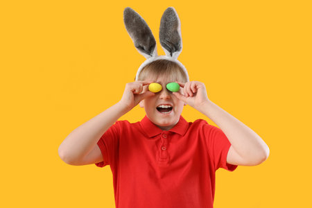 Funny little boy in bunny ears with Easter eggs on yellow background