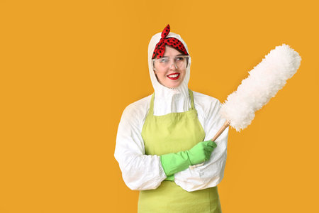 Housewife in protective costume and with dust brush on color background