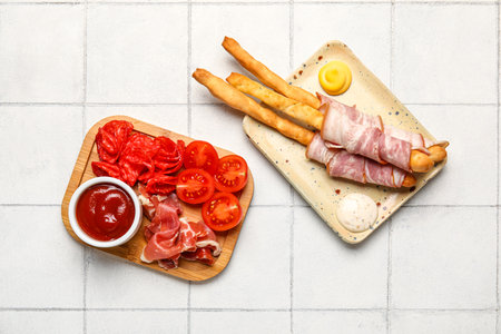 Plate and wooden board of tasty Italian Grissini with bacon on white tile backgroundの写真素材