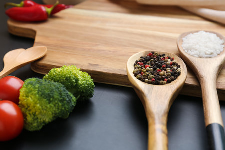 Spoons with spices, fresh vegetables and wooden cutting board on dark background, closeupの写真素材