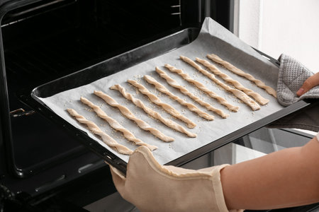 Woman putting baking sheet with Italian Grissini into oven, closeup
