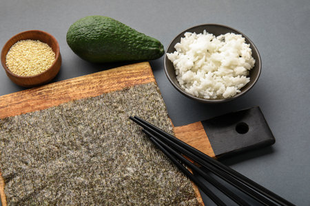 Wooden board with nori sheet, bowl of rice, sesame seeds, avocado and chopsticks on dark background, closeupの写真素材