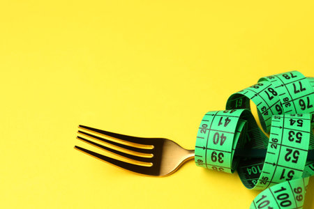 New measuring tape and fork on yellow background, closeup