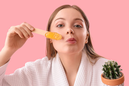 Young woman holding spatula with sugaring paste and cactus on pink backgroundの写真素材