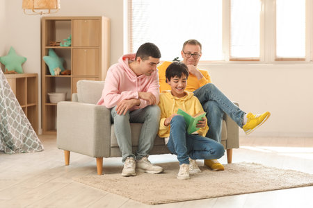 Happy little boy with his dad and grandfather reading book at homeの写真素材
