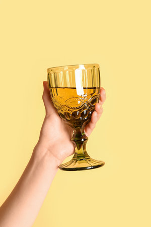 Female hand holding glass of white wine on yellow backgroundの写真素材
