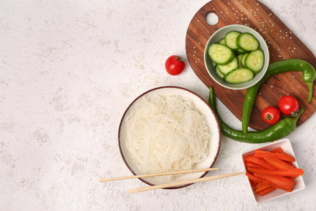 Composition with bowl of tasty rice noodles, chopsticks and vegetables on white grunge tableの写真素材