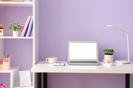 Workplace with laptop cup and stationery near lilac wall in officeの写真素材