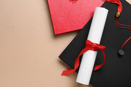 Diploma with red ribbon, graduation hat and book on beige backgroundの写真素材