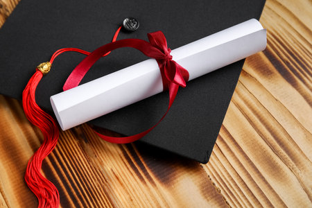 Diploma with red ribbon and graduation hat on wooden tableの写真素材