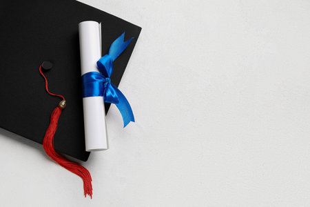 Diploma with blue ribbon and graduation hat on white tableの写真素材