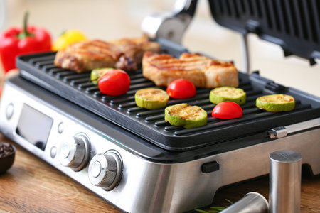 Modern electric grill with tasty steaks and vegetables on tableの写真素材