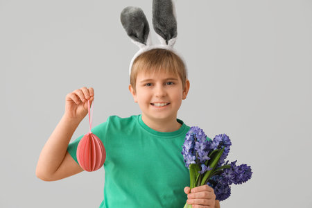 Little boy in bunny ears with paper Easter egg and flowers on gray background