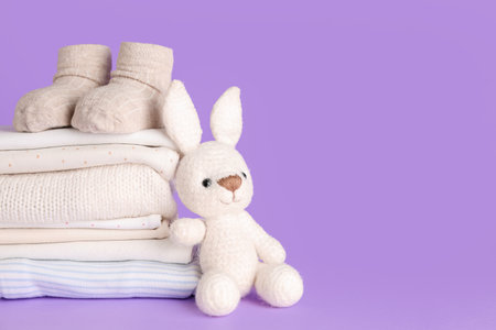Stack of baby clothes, socks and knitted toy on lilac backgroundの写真素材
