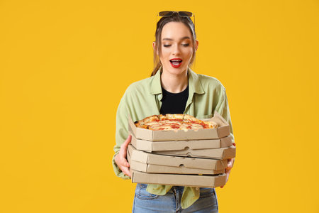 Surprised young woman with cardboard boxes of tasty pizza on yellow backgroundの写真素材