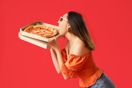Beautiful woman in sunglasses eating tasty pizza on red backgroundの写真素材