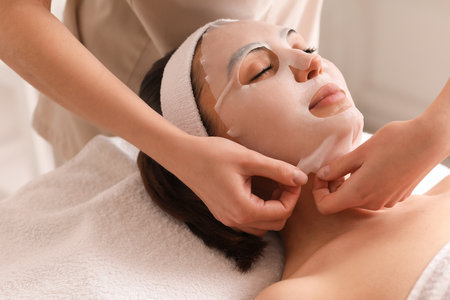 Cosmetologist applying sheet mask onto young woman's face in salon, closeupの写真素材