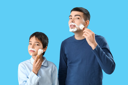Man and his little son applying shaving foam onto faces against blue backgroundの写真素材