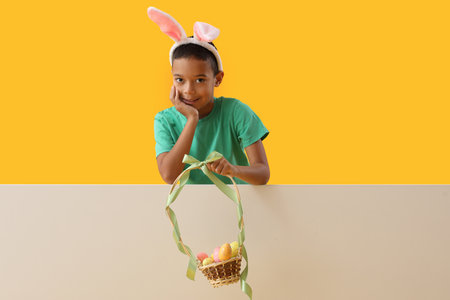 Little African-American boy in bunny ears with basket of Easter eggs on color backgroundの写真素材
