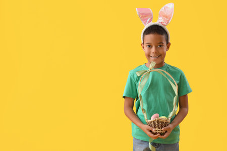 Little African-American boy in bunny ears with basket of Easter eggs on yellow backgroundの写真素材