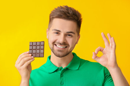 Handsome young man with tasty chocolate showing OK on color backgroundの写真素材