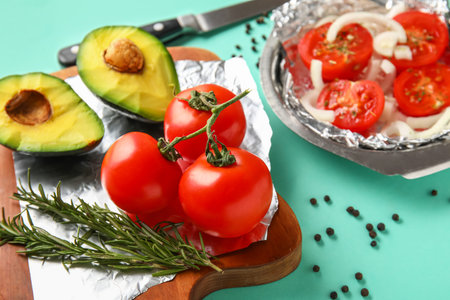 Wooden board with aluminum foil, fresh tomatoes, avocado and rosemary on color background, closeup