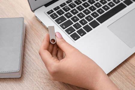 Female hand with USB flash drive, modern laptop and book on wooden table, closeup