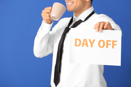 Young businessman holding paper with text DAY OFF and drinking coffee against color backgroundの写真素材