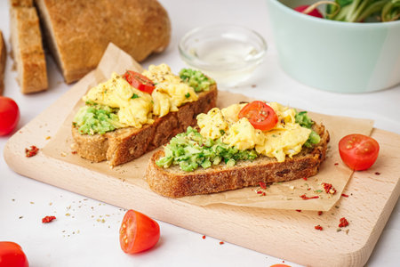 Wooden board with tasty scrambled eggs sandwich and vegetables on light backgroundの写真素材