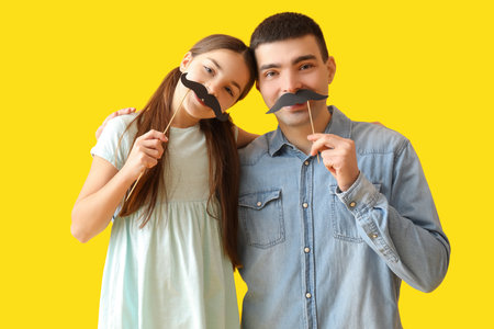 Portrait of father and his little daughter with paper mustache on yellow backgroundの写真素材