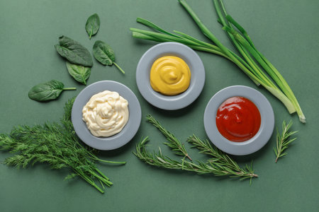 Plates of ketchup, mayonnaise and mustard with herbs on green backgroundの写真素材