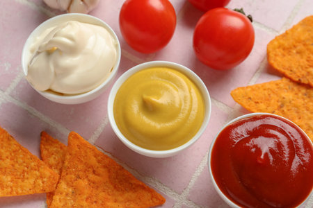 Bowls of ketchup, mayonnaise and mustard with nachos and tomatoes on pink tiled table